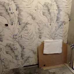 Ściana łazienki w trakcie układania płytek z motywem liści, widoczne krzyżyki dystansowe i przygotowane płyty do dalszych prac.