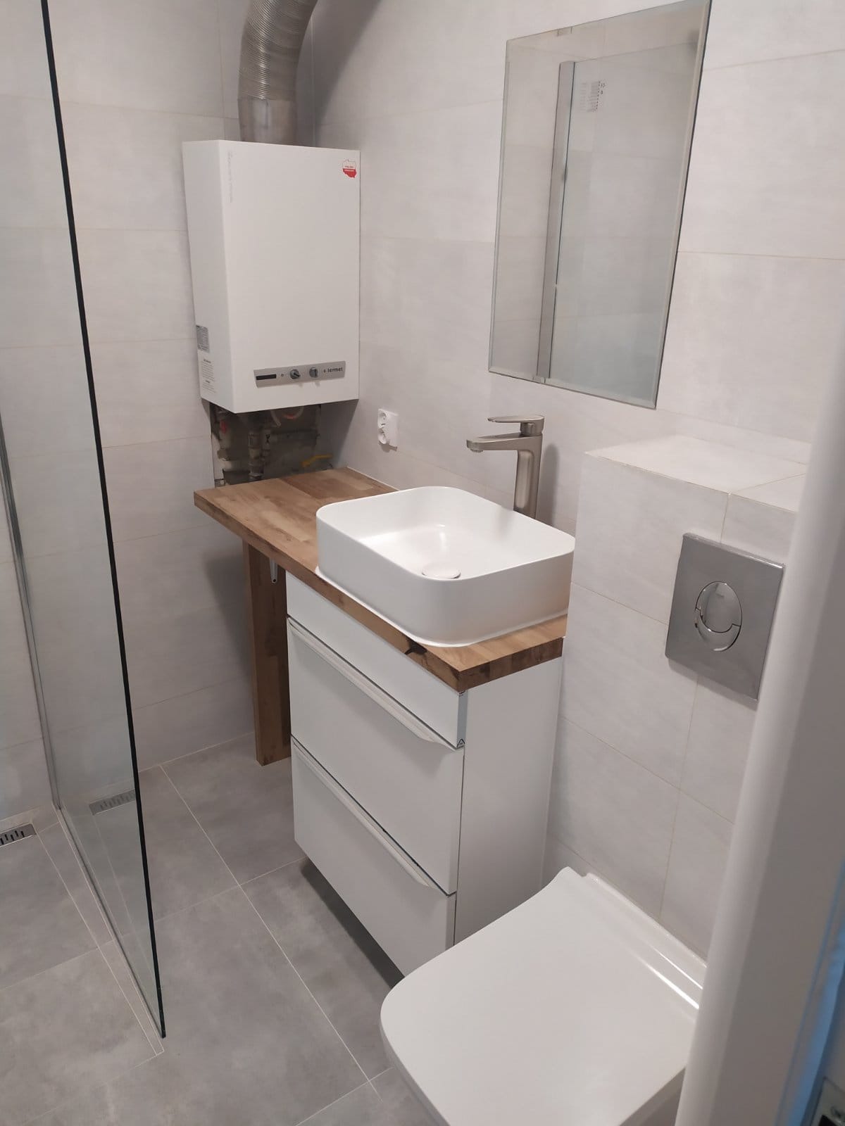 Nowoczesna łazienka z białą umywalką nablatową na szafce z drewnianym blatem, wiszącą toaletą i piecem gazowym, całość w jasnych płytkach.