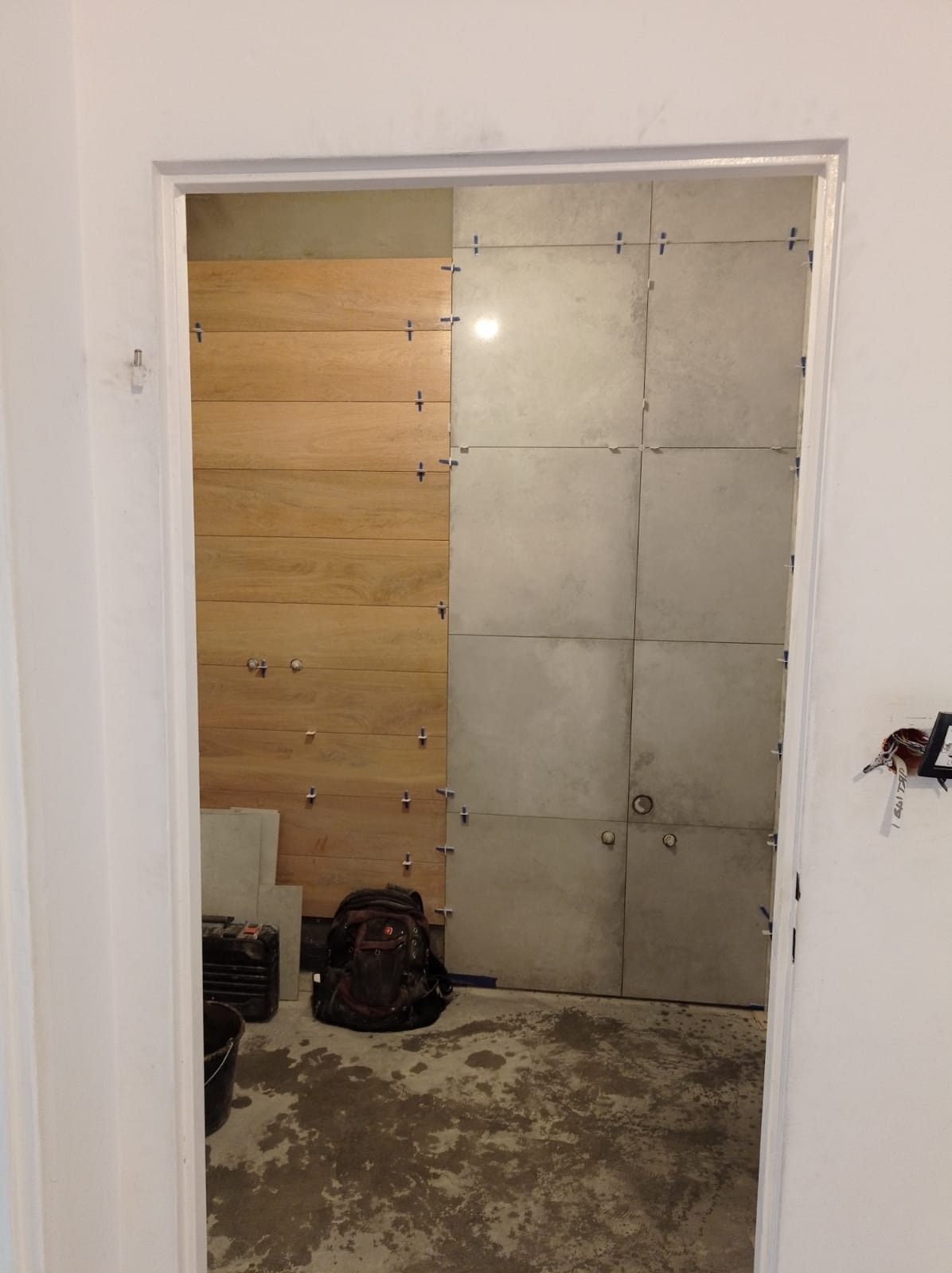 Ściana w trakcie układania płytek: po lewej drewnopodobne panele, po prawej szare płytki wielkoformatowe z krzyżykami dystansowymi, widoczny plecak i narzędzia na podłodze.