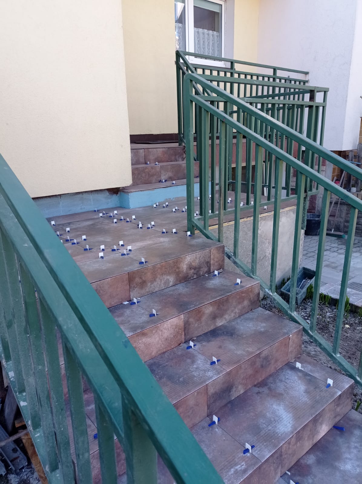 Układanie brązowych płytek na schodach wejściowych, widoczne plastikowe krzyżyki dystansowe, zielona metalowa balustrada.