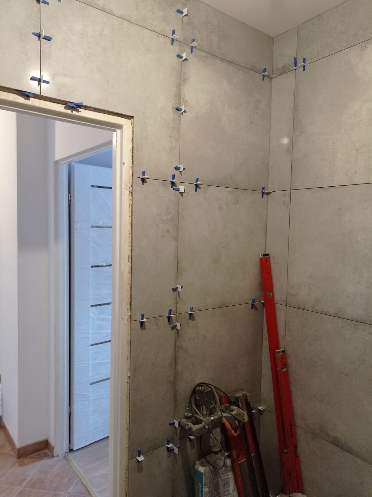 Świeżo ułożone szare płytki na ścianie łazienki, z widocznymi krzyżykami dystansowymi i poziomnicą opartą o ścianę, obok widać narzędzia glazurnicze i fragment drzwi.