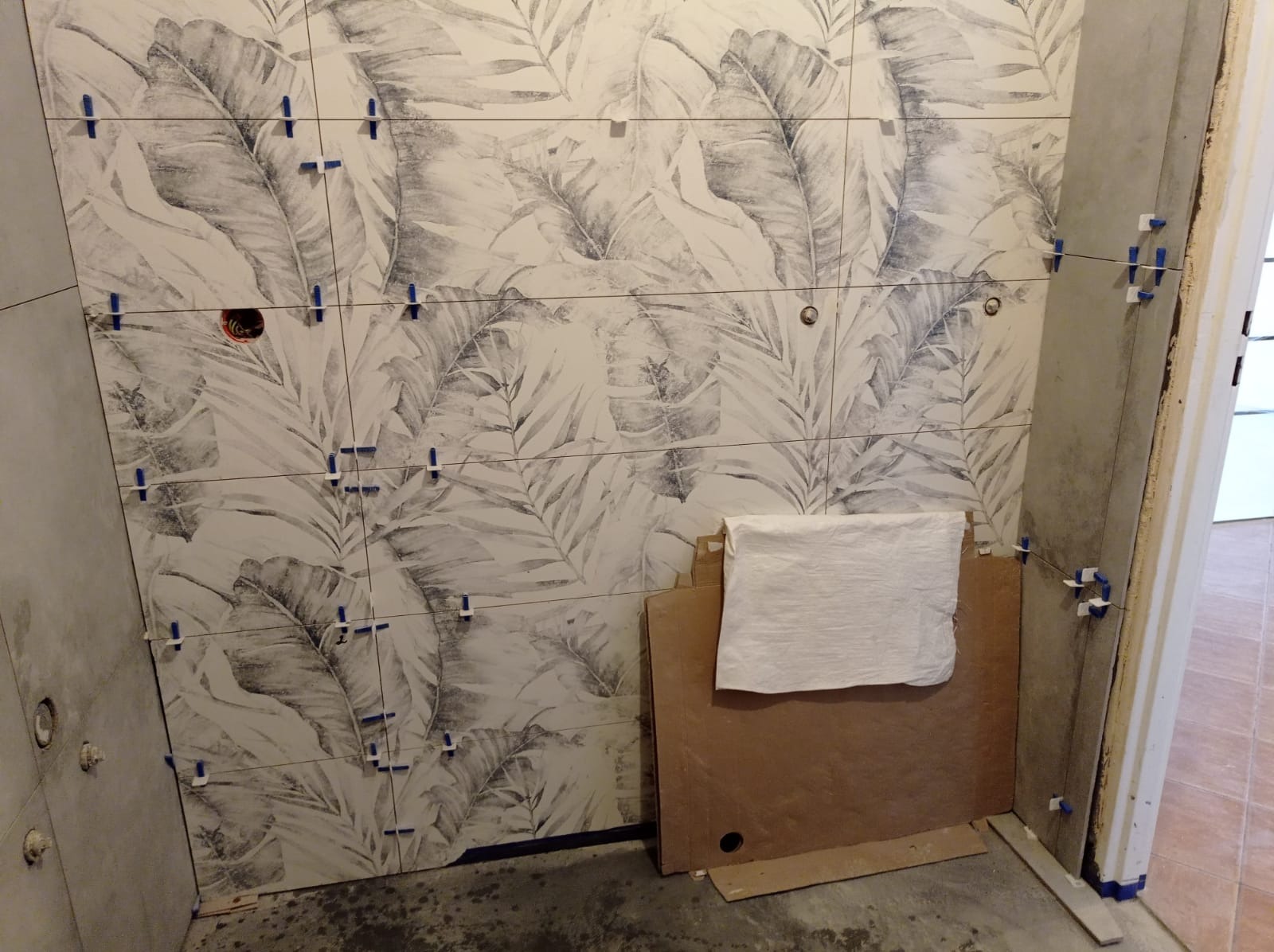 Ściana łazienki w trakcie układania płytek z motywem liści, widoczne krzyżyki dystansowe i przygotowane płyty do dalszych prac.