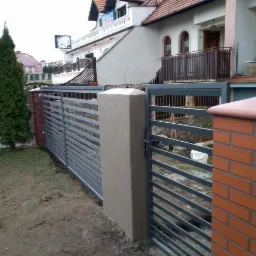 Nowoczesne ogrodzenie posesji z szarych, poziomych przęseł, osadzone na słupkach z cegły klinkierowej i betonu architektonicznego, widoczne na tle domu jednorodzinnego.