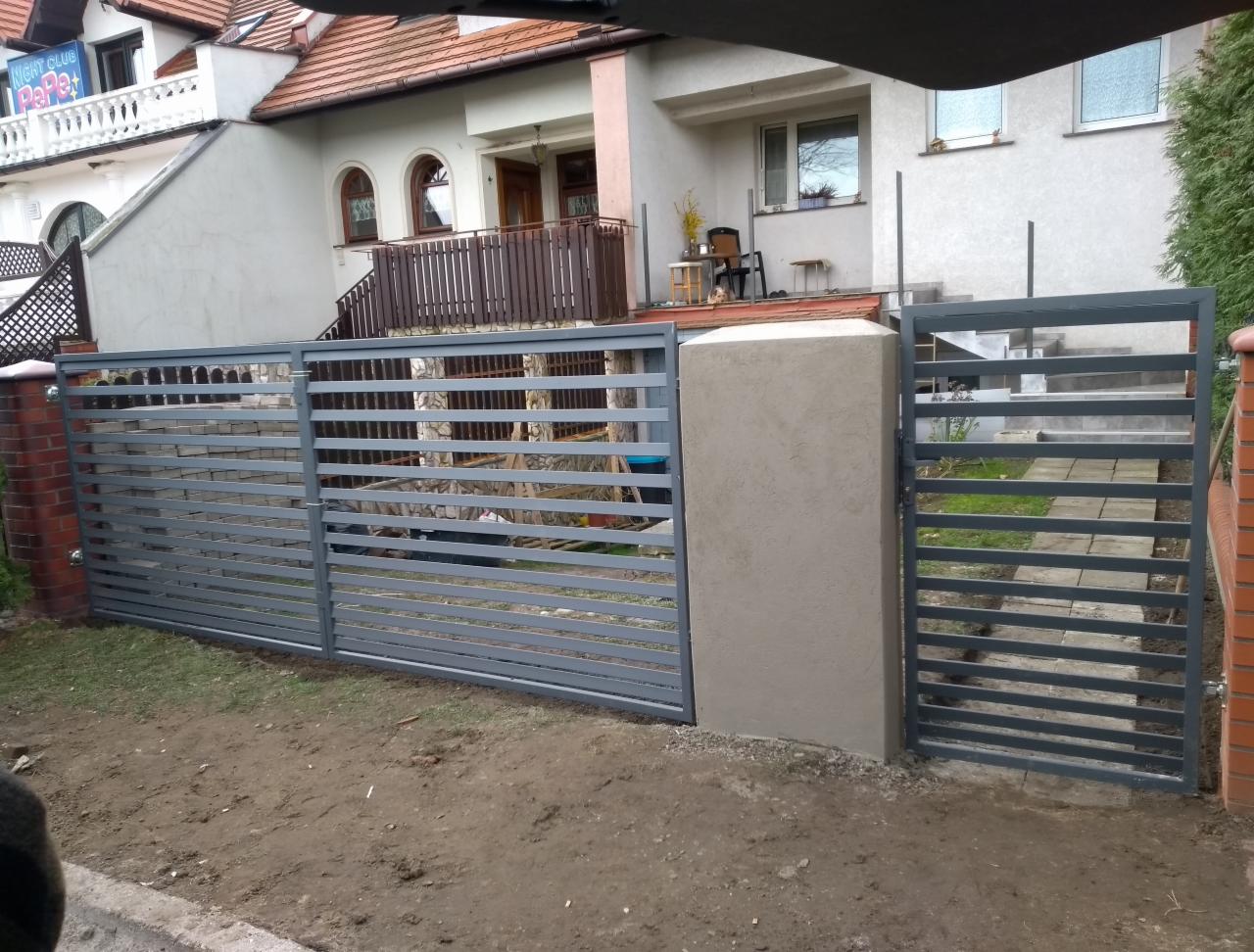 Nowoczesne, poziome ogrodzenie z szarych profili stalowych, zamontowane między murowanymi słupkami z cegły klinkierowej i betonowym słupem, na tle dwukondygnacyjnego domu z beżową elewacją.