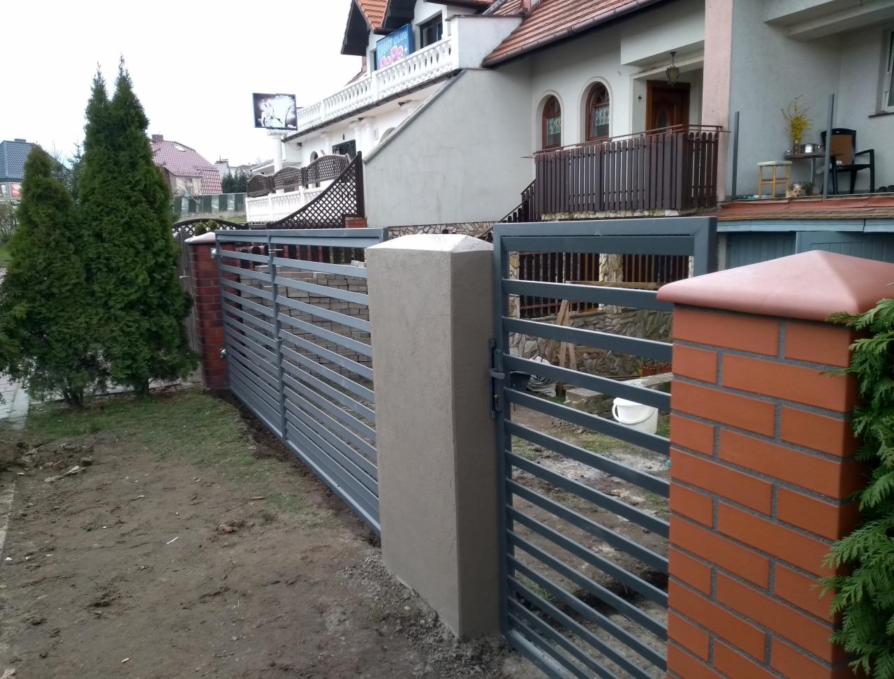 Nowoczesne ogrodzenie posesji z szarych, poziomych przęseł, osadzone na słupkach z cegły klinkierowej i betonu architektonicznego, widoczne na tle domu jednorodzinnego.