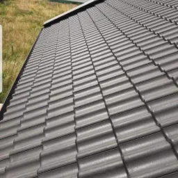 Nowy dach z ciemnoszarej dachówki ceramicznej, widok z góry, w tle budynek mieszkalny i pola uprawne.