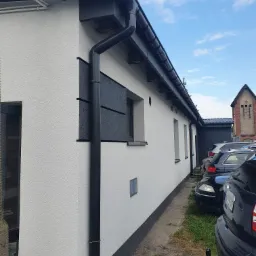 Boczny widok budynku z białą elewacją o strukturze baranka, czarną rynną i fragmentem czarnej, płaskiej okładziny elewacyjnej, na tle zaparkowanych samochodów i budynku z czerwonej cegły.