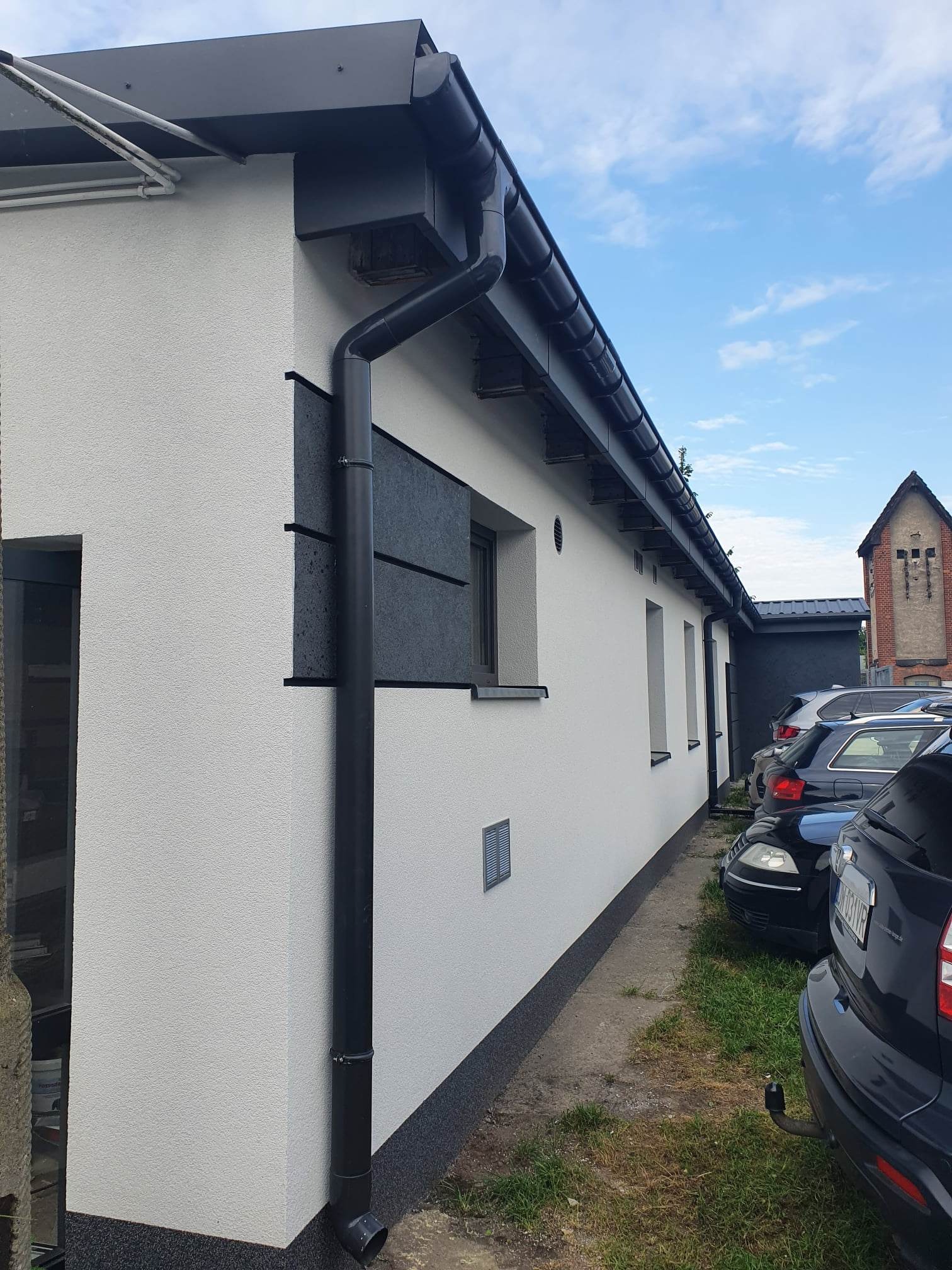 Boczny widok budynku z białą elewacją o strukturze baranka, czarną rynną i fragmentem czarnej, płaskiej okładziny elewacyjnej, na tle zaparkowanych samochodów i budynku z czerwonej cegły.