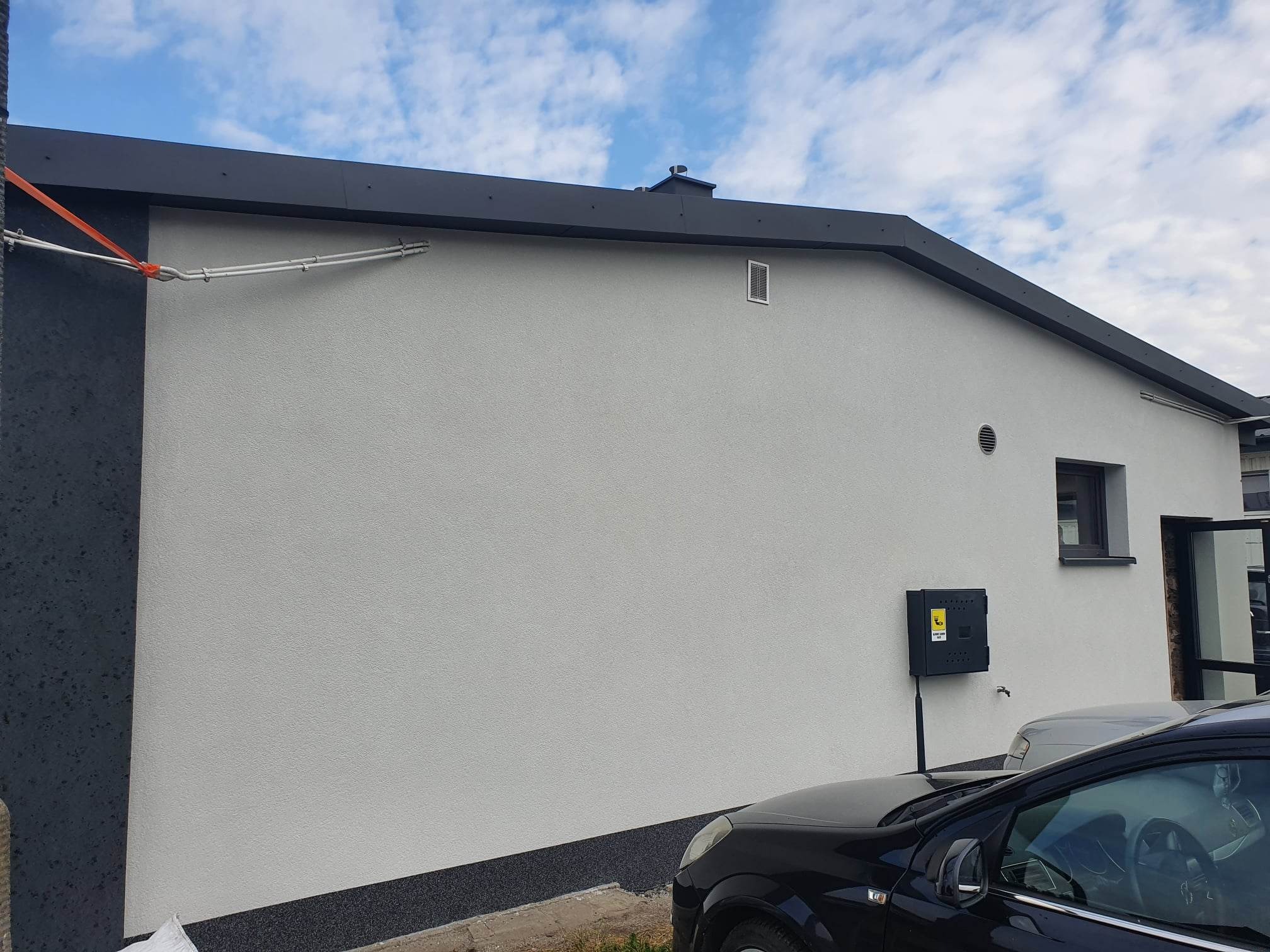 Jasnoszara elewacja domu jednorodzinnego z czarnym obramowaniem dachu, widoczna skrzynka elektryczna na ścianie i fragment zaparkowanego samochodu.