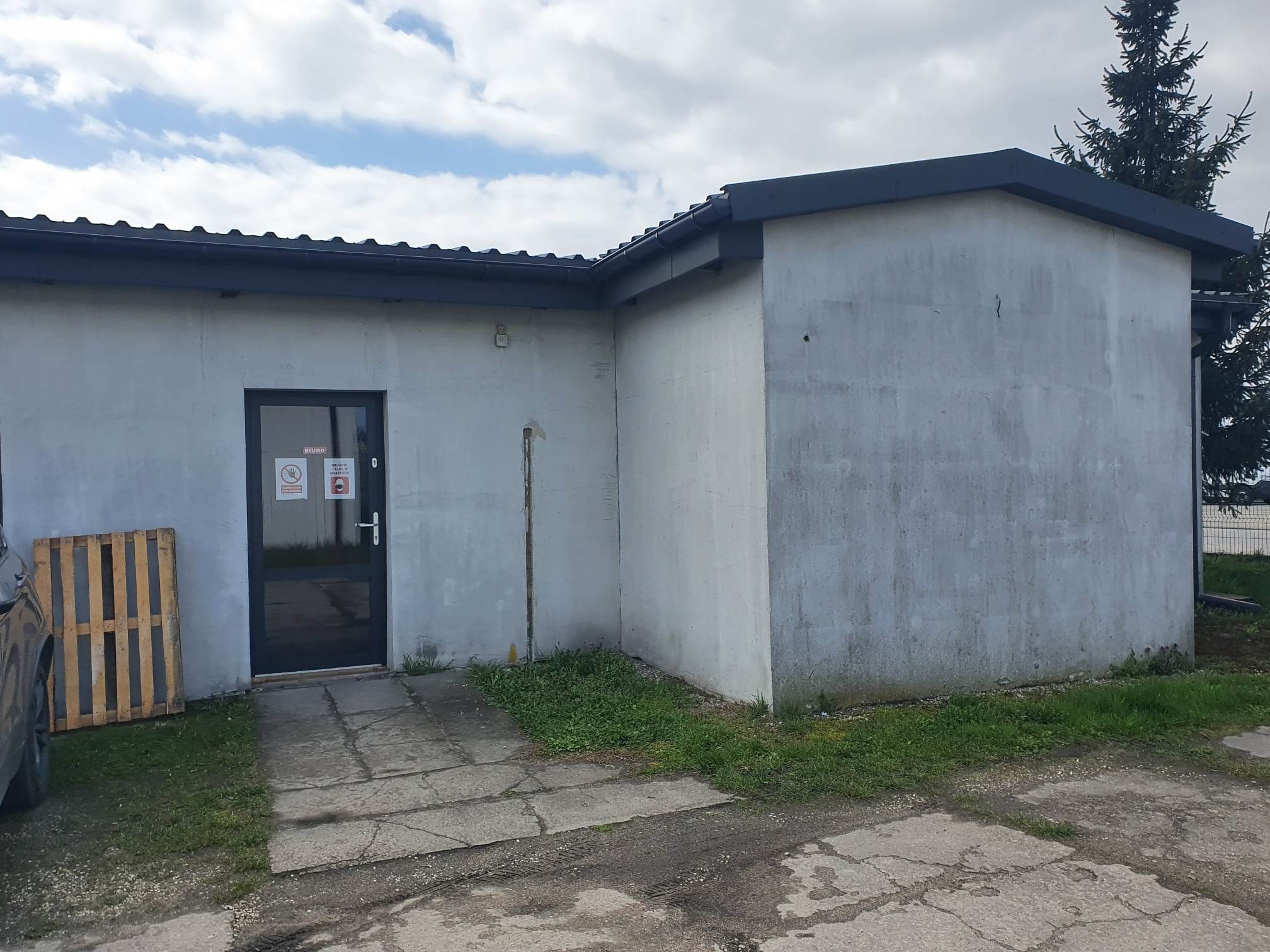 Szara elewacja budynku z ciemnymi obramowaniami okien i dachu, widoczna paleta drewniana obok drzwi wejściowych, zarośnięty trawnik u podstawy elewacji.