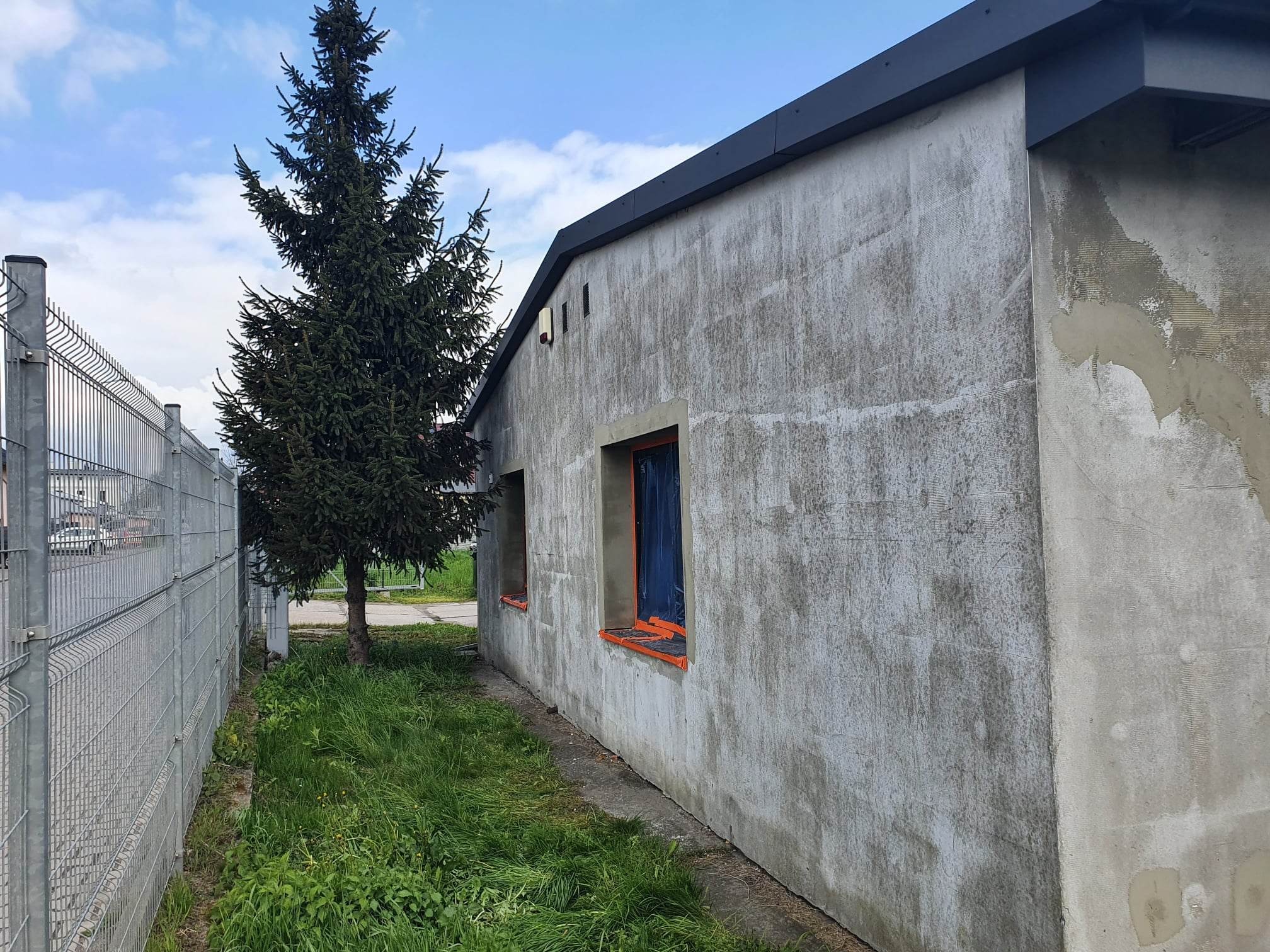 Budynek w trakcie przygotowania do elewacji, okna zabezpieczone folią, widoczne ślady wcześniejszych prac, szara elewacja w surowym stanie, obok rośnie świerk i znajduje się metalowe ogrodzenie.