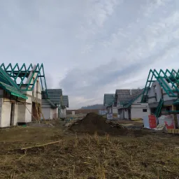 Szereg surowych, murowanych domów w trakcie budowy z widoczną konstrukcją dachu z zielonych belek, białe ściany z bloczków, sterta ziemi na pierwszym planie i pochmurne niebo w tle.