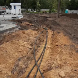 Wykop w ziemi z ułożonymi czarnymi rurami, widoczne narzędzia i biały kontener w tle, sugerujące prace instalacyjne w ogrodzie lub na działce.