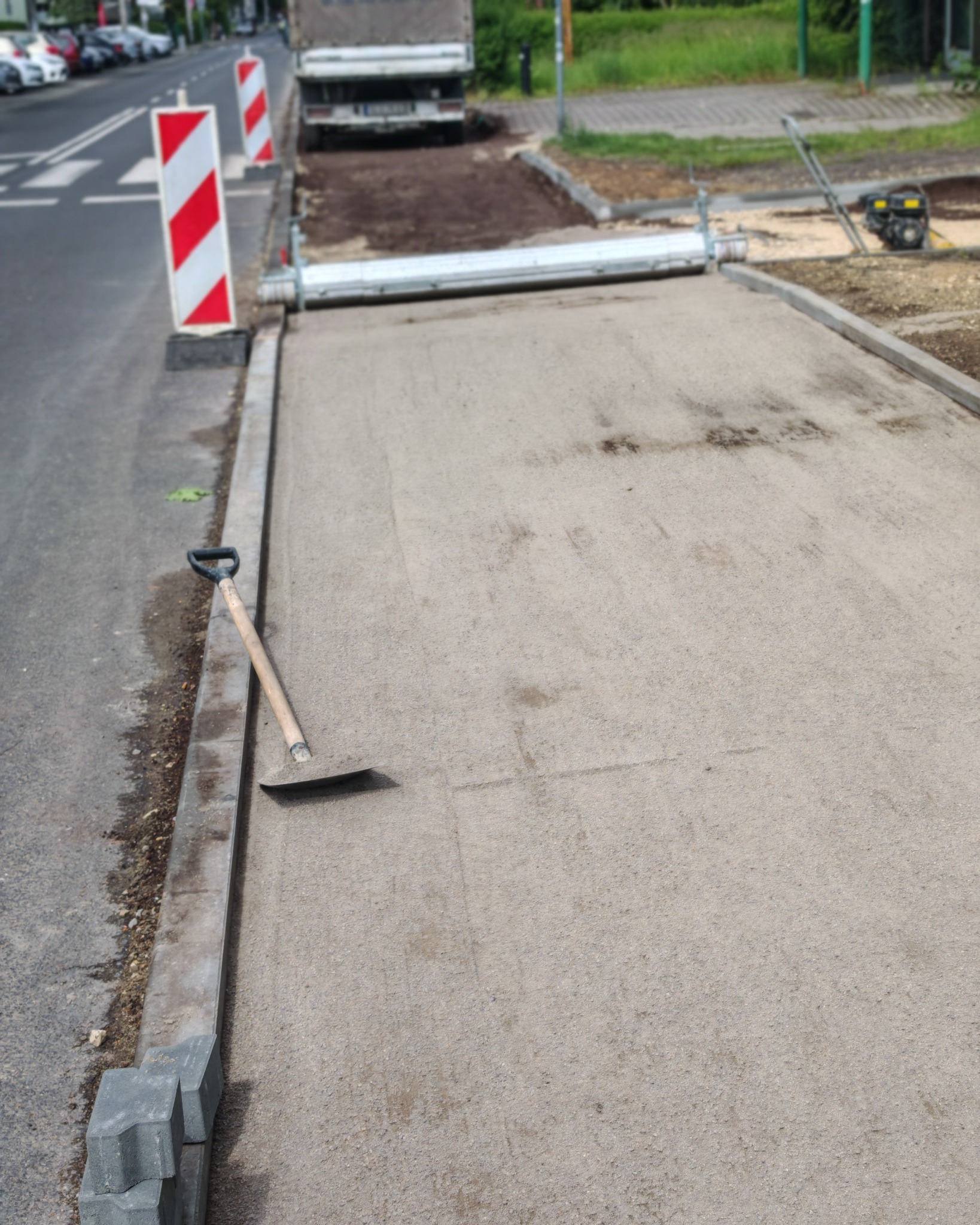 Przygotowany teren pod kostkę brukową z narzędziami i częściowo ułożonym obrzeżem. Widoczne znaki ostrzegawcze i sprzęt budowlany w tle. Etap prac.