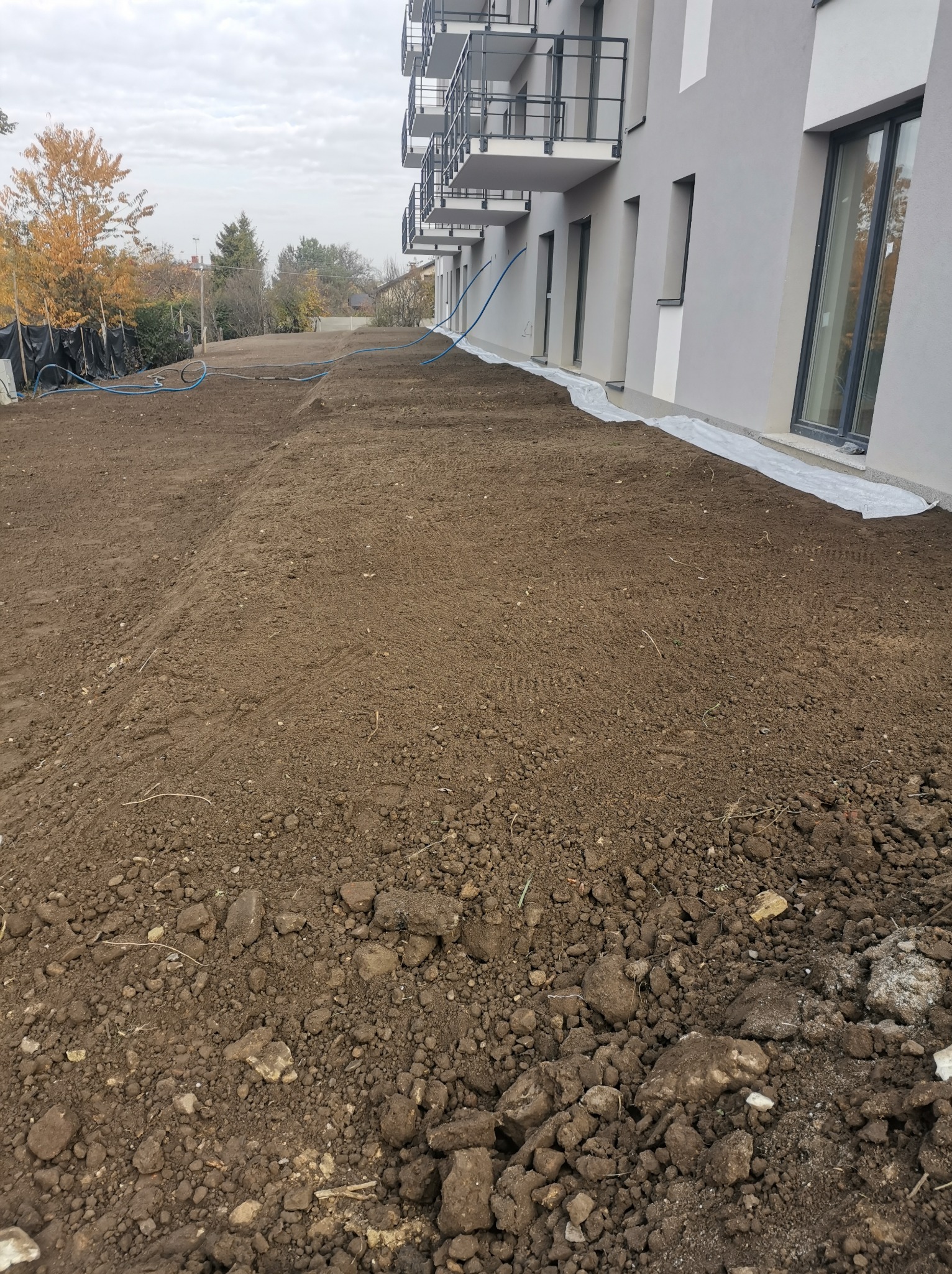 Przygotowany teren pod budowę z widoczną warstwą ziemi, zabezpieczeniem budynku folią i niebieskimi rurami. W tle drzewa i budynki.