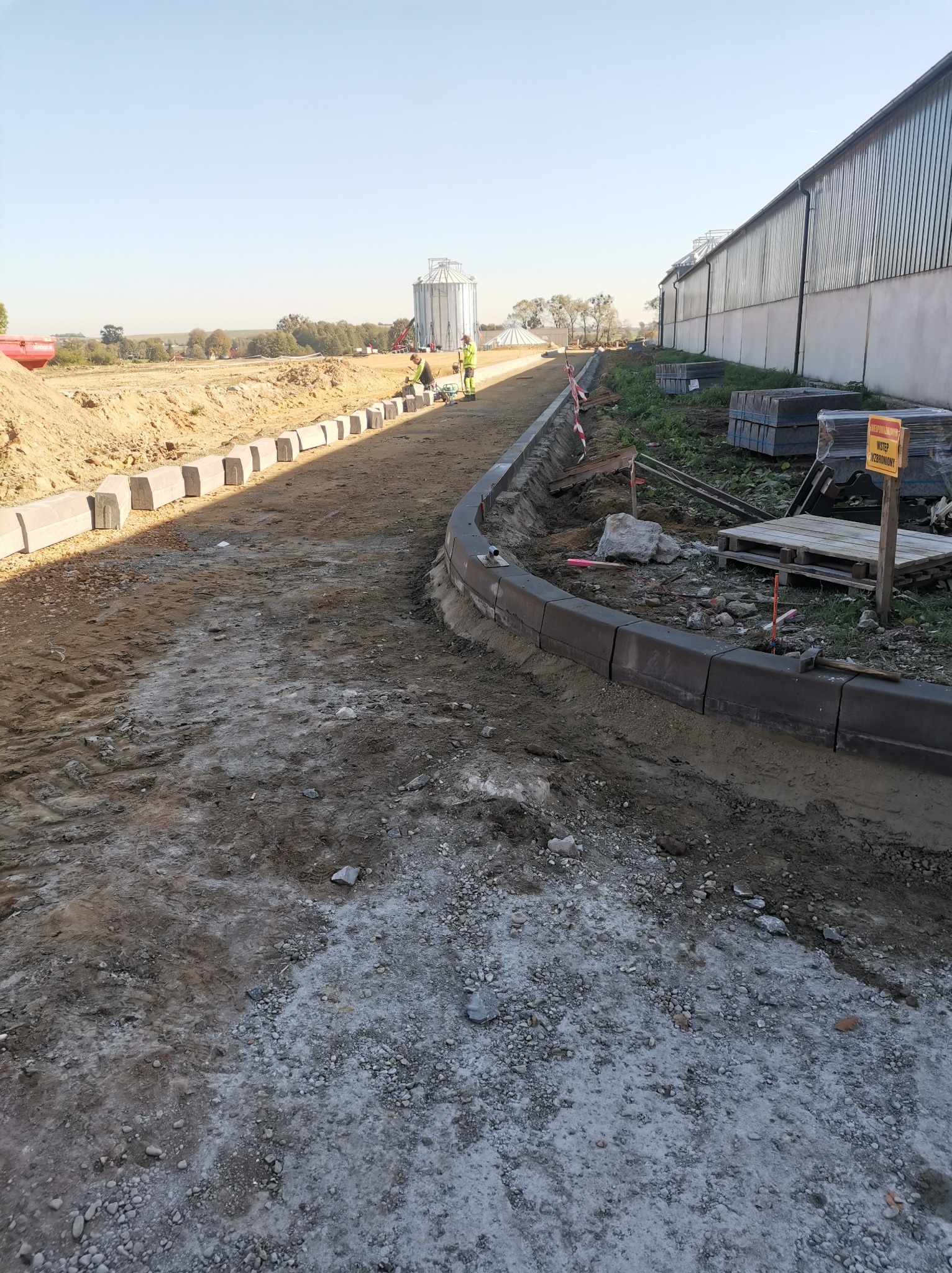 Montaż obrzeży betonowych przy drodze gruntowej. W tle budynek gospodarczy i pracownicy w odblaskowych ubraniach. Widoczny znak ostrzegawczy i ułożone palety z kostką.