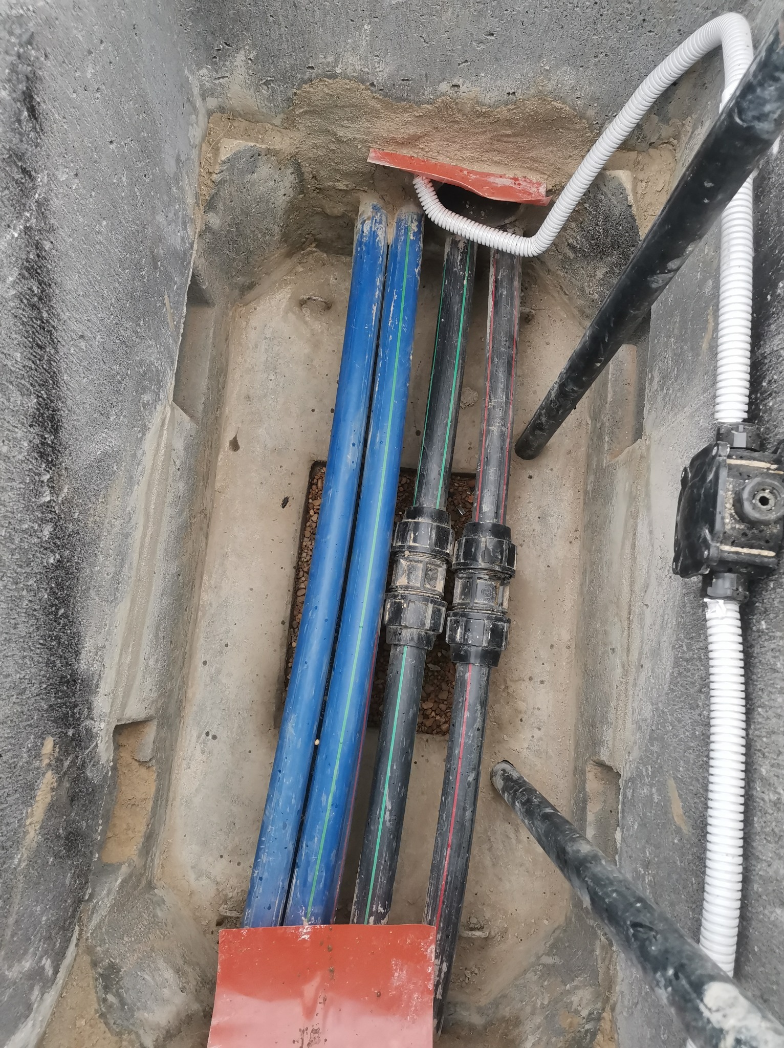 Wykop z ułożonymi rurami: niebieskie, czarne z zielonym paskiem, biała karbowana i czarna, sztywna. Widoczne złączki i elementy instalacji podziemnej.