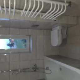 Wykończona łazienka z beżowymi płytkami, widoczna wanna, toaleta podwieszana i grzejnik drabinkowy na ścianie, z oknem z widokiem na zewnątrz.