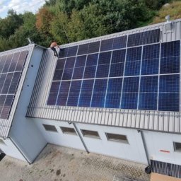 IET-GAWRYCH - Instalacja fotowoltaiczna na dachu budynku z blachy trapezowej, widok z góry. Jeden z instalatorów pracuje na dachu. Część paneli jeszcze nie zamontowana.