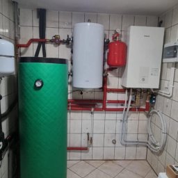 IET-GAWRYCH - Zestaw instalacji grzewczej w kotłowni: zielony zbiornik buforowy, biały zasobnik CWU, czerwony zbiornik przeponowy i wiszący piec grzewczy. Widoczne rury i armatura.