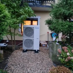 IET-GAWRYCH - Biała pompa ciepła Panasonic zainstalowana na żwirowym podłożu przed budynkiem z balkonem, otoczona zielenią. Widoczne narzędzia i karton po instalacji.