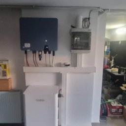 IET-GAWRYCH - Kompletna instalacja fotowoltaiczna w pomieszczeniu: inwerter na ścianie, obok skrzynka elektryczna, na dole akumulator. Widoczne przewody i elementy montażowe.