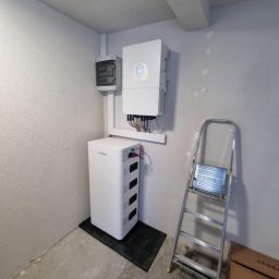 IET-GAWRYCH - Biały akumulator energii, inwerter i skrzynka elektryczna zamontowane na szarej ścianie w pomieszczeniu. Aluminiowa drabina oparta o ścianę. Widok z dołu.