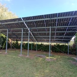 IET-GAWRYCH - Montaż paneli słonecznych na metalowej konstrukcji w ogrodzie. Widoczna osoba z drabiną i częściowo zamontowane panele. Ujęcie z dołu, na tle zieleni i błękitnego nieba.