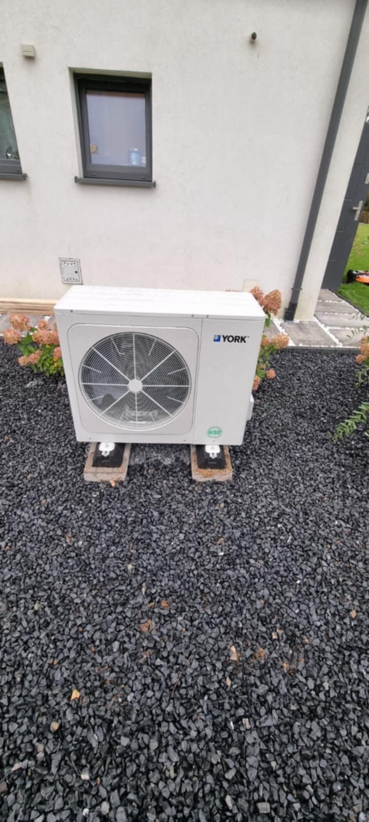 Biała pompa ciepła York na tle elewacji domu i czarnego żwiru. Widoczne kwiaty hortensji i fragment trawnika. Ujęcie z poziomu gruntu.