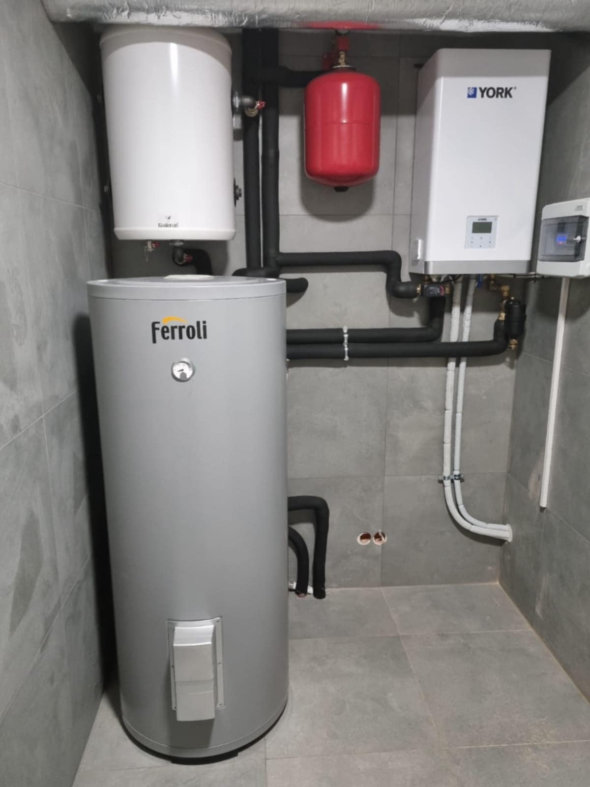 Zestaw urządzeń grzewczych: szary zasobnik Ferroli, biały zasobnik Galmet, biały piec York i czerwony zbiornik wyrównawczy, połączone czarnymi rurami w kotłowni.