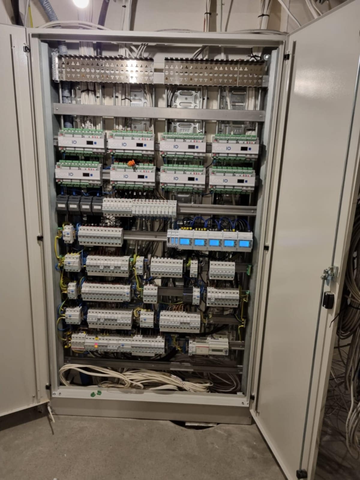 Otwarta szafa sterownicza z licznymi modułami, przewodami i złączkami. Widoczne elementy automatyki i zabezpieczeń. Instalacja elektryczna w szarej obudowie.