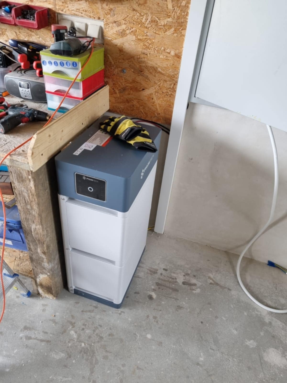 Biało-szara bateria magazynu energii Pylontech, z żółto-czarnymi rękawicami roboczymi na górze, stoi w rogu pomieszczenia z boazerią i betonową podłogą.