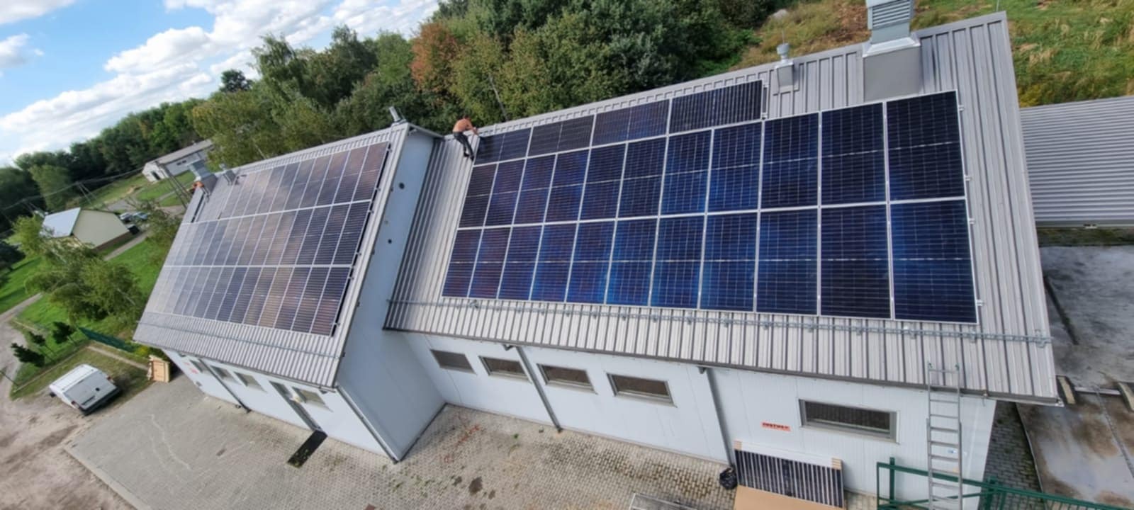 Instalacja fotowoltaiczna na dachu budynku z blachy trapezowej, widok z góry. Jeden z instalatorów pracuje na dachu. Część paneli jeszcze nie zamontowana.