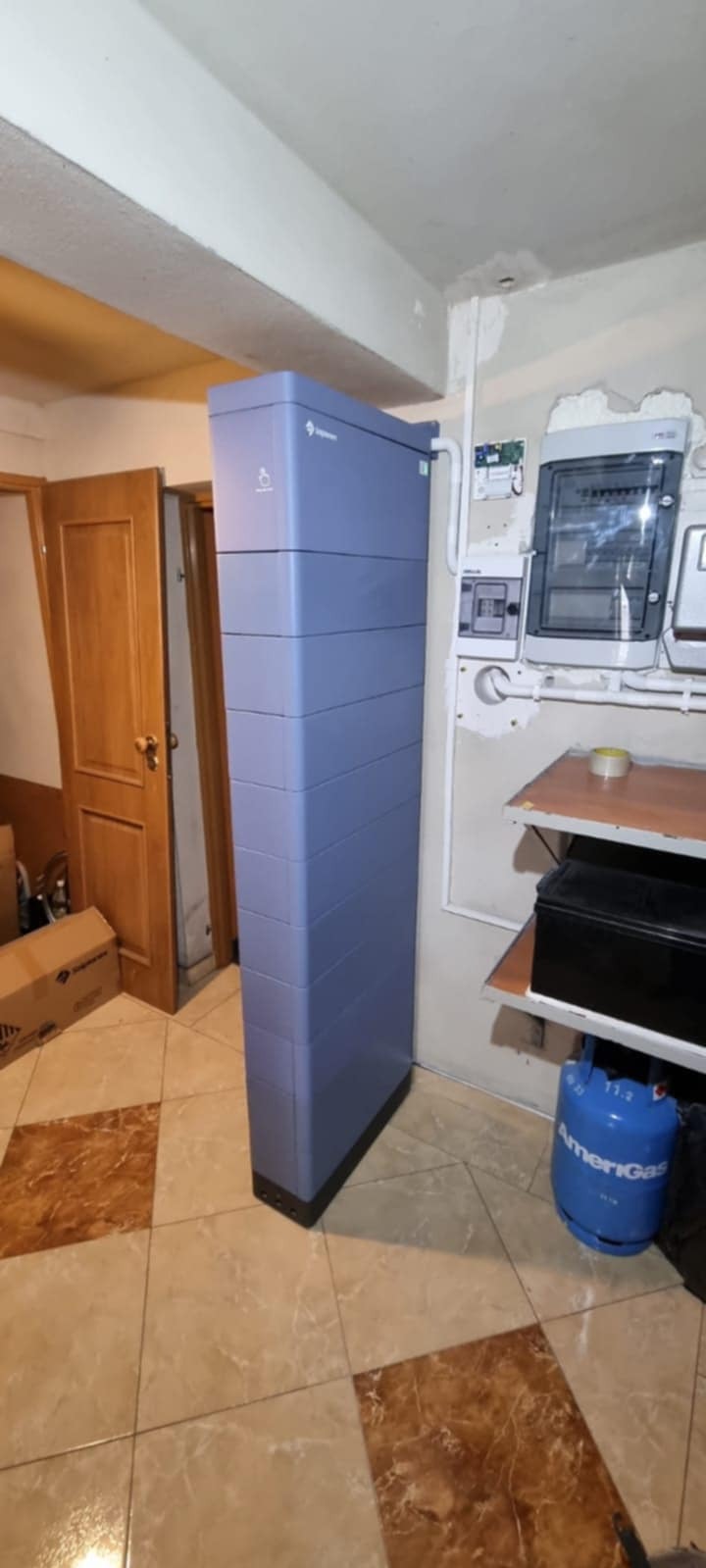 Błękitny akumulator energii Ingeteam stojący w rogu pomieszczenia z brązowymi drzwiami i płytkami podłogowymi. Widoczna instalacja elektryczna na ścianie.