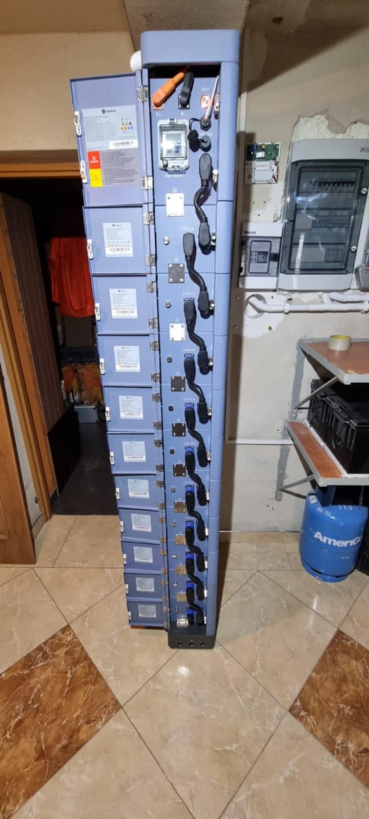 Rozdzielnica elektryczna z otwartymi drzwiczkami, widoczne kable i elementy instalacji. Obok ściana z inną rozdzielnicą i butla gazowa. Ujęcie z dołu.