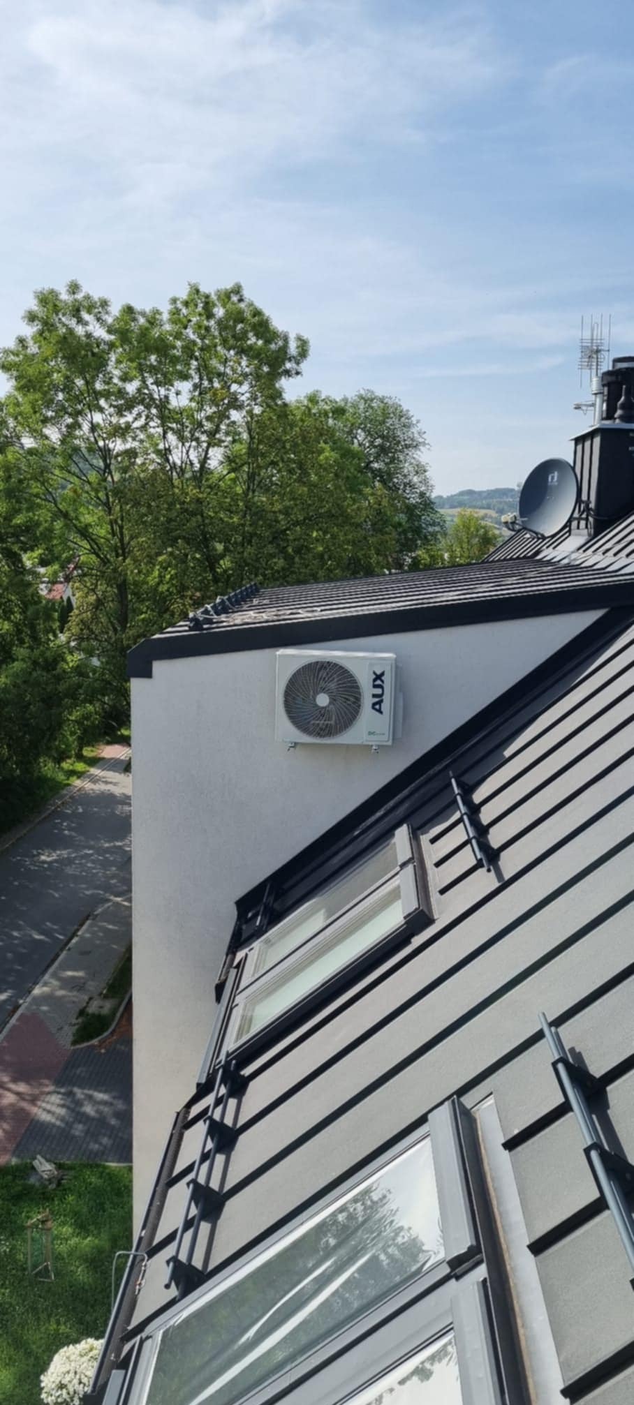 Biała jednostka zewnętrzna klimatyzacji AUX zamontowana na ścianie domu z czarnym dachem i oknami dachowymi. Widok z góry, drzewa w tle.