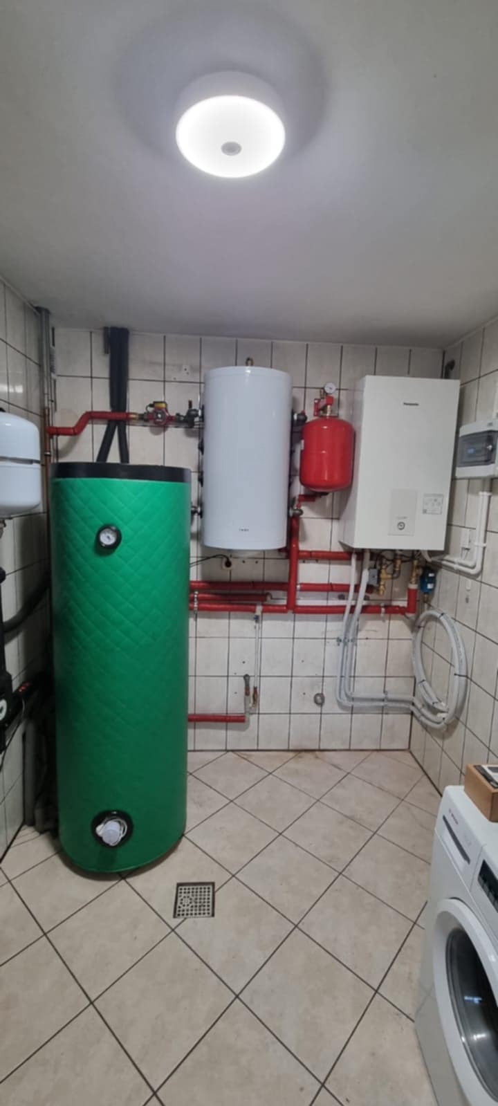 Zestaw instalacji grzewczej w kotłowni: zielony zbiornik buforowy, biały zasobnik CWU, czerwony zbiornik przeponowy i wiszący piec grzewczy. Widoczne rury i armatura.