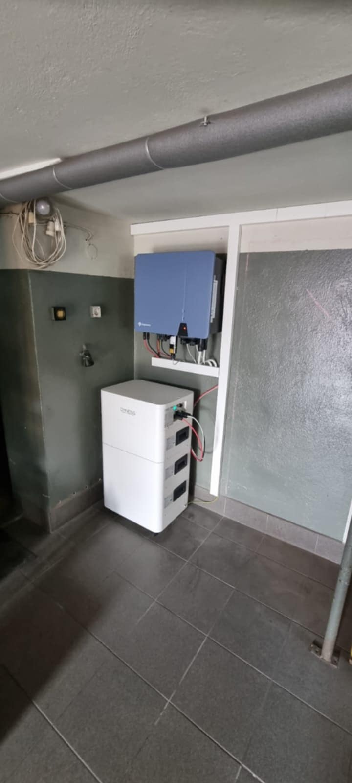 Biały akumulator energii i niebieski inwerter fotowoltaiczny zamontowane w pomieszczeniu z szarymi płytkami podłogowymi i rurą pod sufitem.