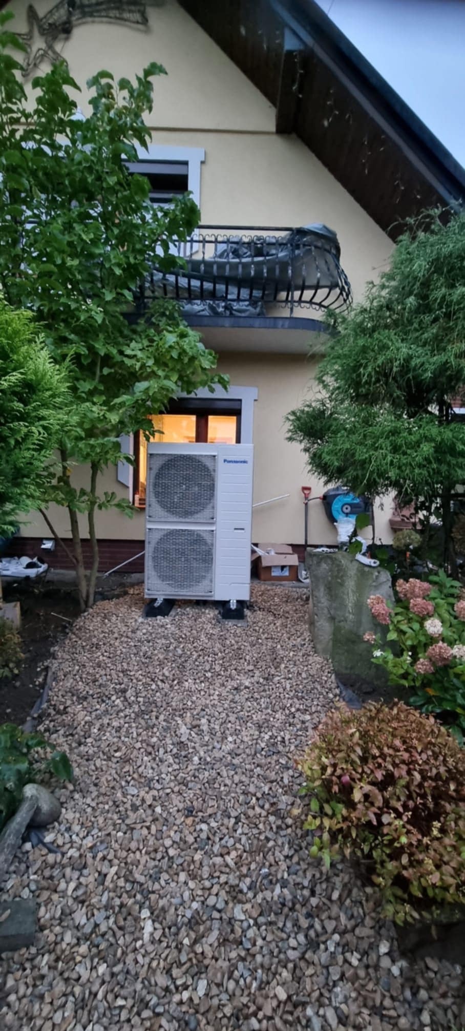 Biała pompa ciepła Panasonic zainstalowana na żwirowym podłożu przed budynkiem z balkonem, otoczona zielenią. Widoczne narzędzia i karton po instalacji.