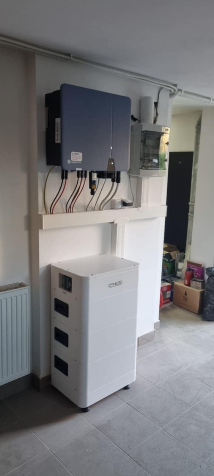 Biały akumulator energii Dyness obok szarej skrzynki z kablami na ścianie w pomieszczeniu. Widoczne kable, rury i grzejnik. Szare płytki na podłodze.