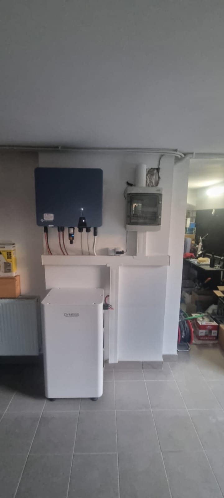 Kompletna instalacja fotowoltaiczna w pomieszczeniu: inwerter na ścianie, obok skrzynka elektryczna, na dole akumulator. Widoczne przewody i elementy montażowe.