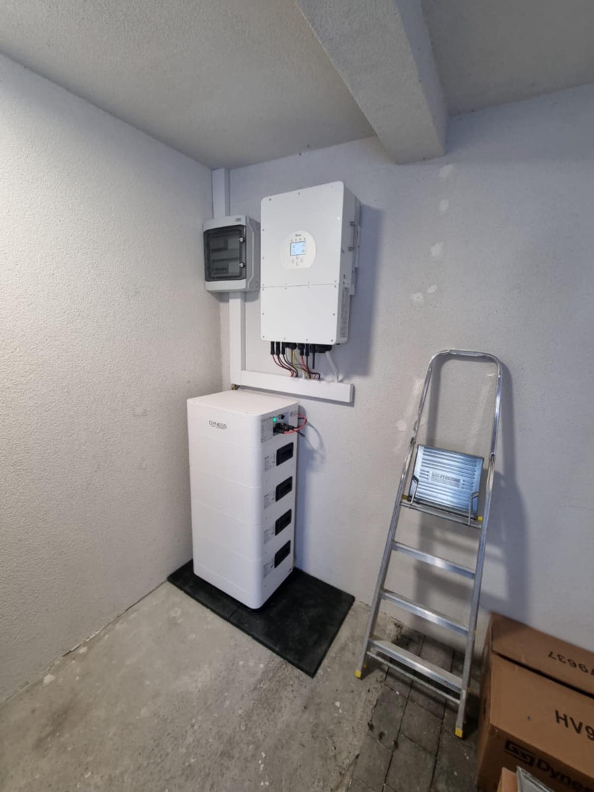 Biały akumulator energii, inwerter i skrzynka elektryczna zamontowane na szarej ścianie w pomieszczeniu. Aluminiowa drabina oparta o ścianę. Widok z dołu.