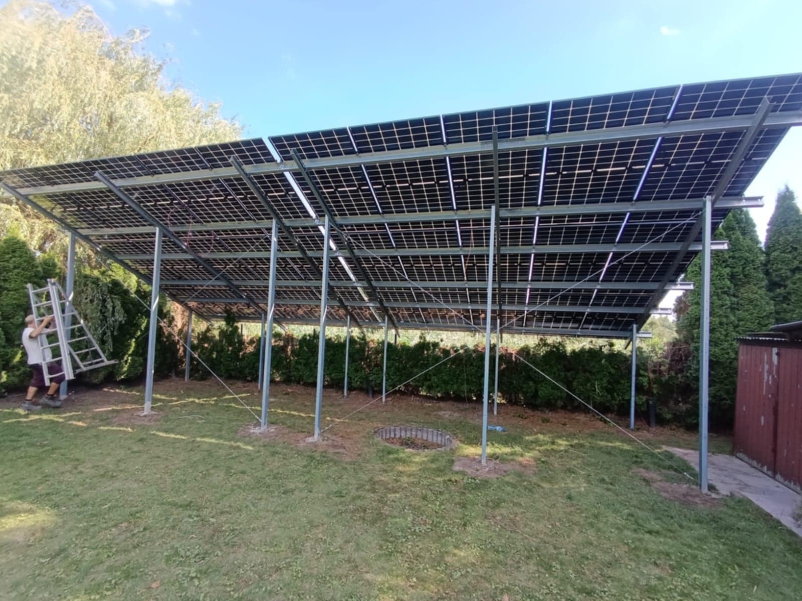 Montaż paneli słonecznych na metalowej konstrukcji w ogrodzie. Widoczna osoba z drabiną i częściowo zamontowane panele. Ujęcie z dołu, na tle zieleni i błękitnego nieba.