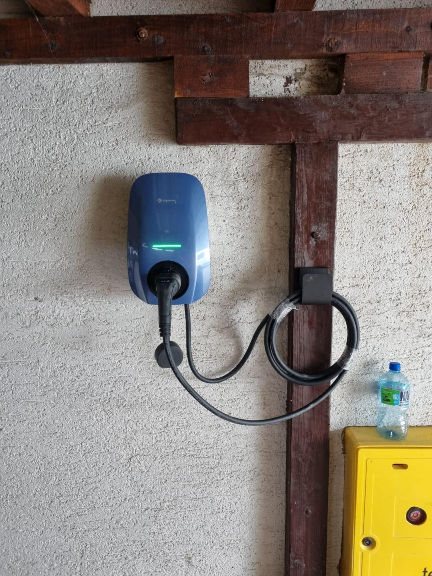 Niebieska stacja ładowania pojazdów elektrycznych zamontowana na szarej, teksturowanej ścianie, z widocznym kablem i butelką wody obok żółtej skrzynki.