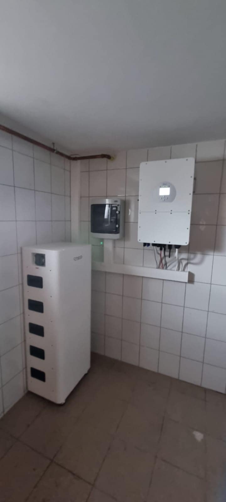 Biały akumulator energii, inwerter i licznik na ścianie w pomieszczeniu z kafelkami. Instalacja fotowoltaiczna wewnątrz budynku.
