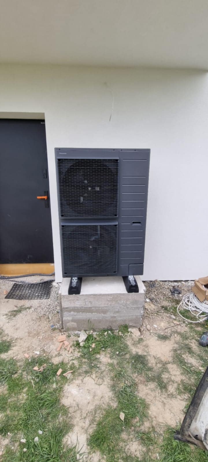 Szara pompa ciepła powietrze-woda, zamontowana na betonowym fundamencie przy białej ścianie budynku z ciemnymi drzwiami. Widoczne przewody i fragment trawnika.