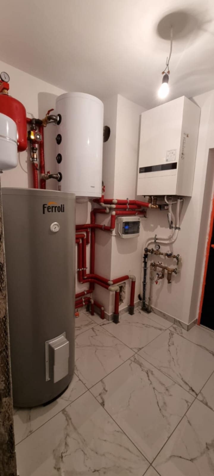 Kompletna instalacja grzewcza z kotłem Ferroli, zasobnikiem i osprzętem, z czerwonymi rurami, na tle białych ścian i marmurowej podłogi. Widok na całą kotłownię.