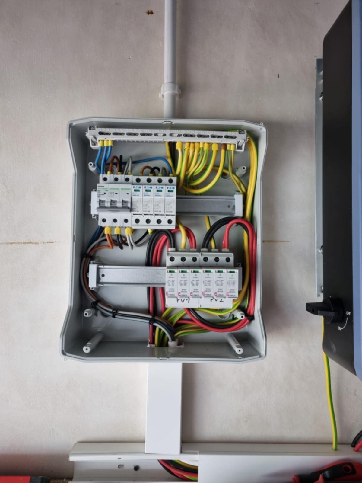 Rozdzielnica elektryczna z zabezpieczeniami przepięciowymi, okablowaniem w kolorach, zamontowana na ścianie. Widoczny fragment falownika.