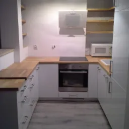 Nowoczesna, jasna kuchnia z drewnianym blatem, białymi szafkami z szufladami i wbudowanym piekarnikiem, oświetlona lampą sufitową i dodatkowym oświetleniem podszafkowym.