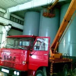 Czerwony samochód ciężarowy STAR z podnośnikiem koszowym na tle dużych, szarych zbiorników w hali przemysłowej; otwarte drzwi kabiny kierowcy.